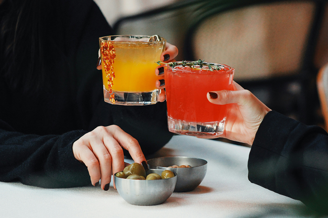 Two women, each holding a different cocktail, snacking on olives and nuts