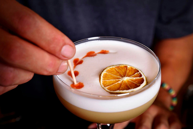 Close up of a cocktail with a thick foam top and dehydrated citrus garnish. A hand holding a cocktail stick adds a final colorful flourish