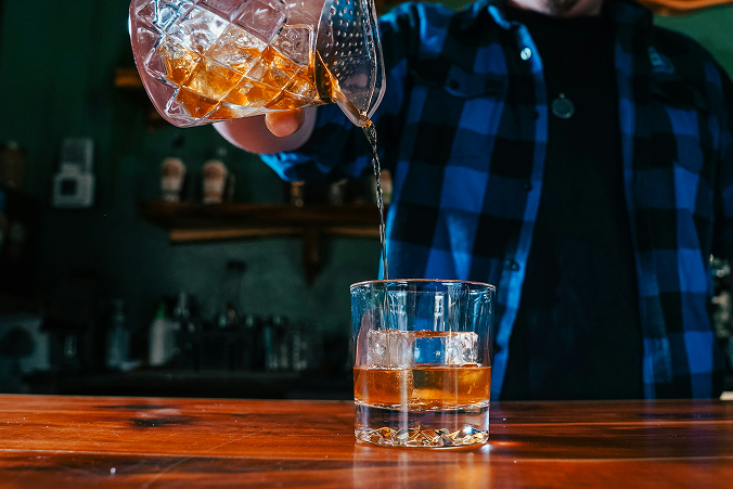A man in a blue flannel shirt pours a cocktail from a mixing glass into a rocks glass